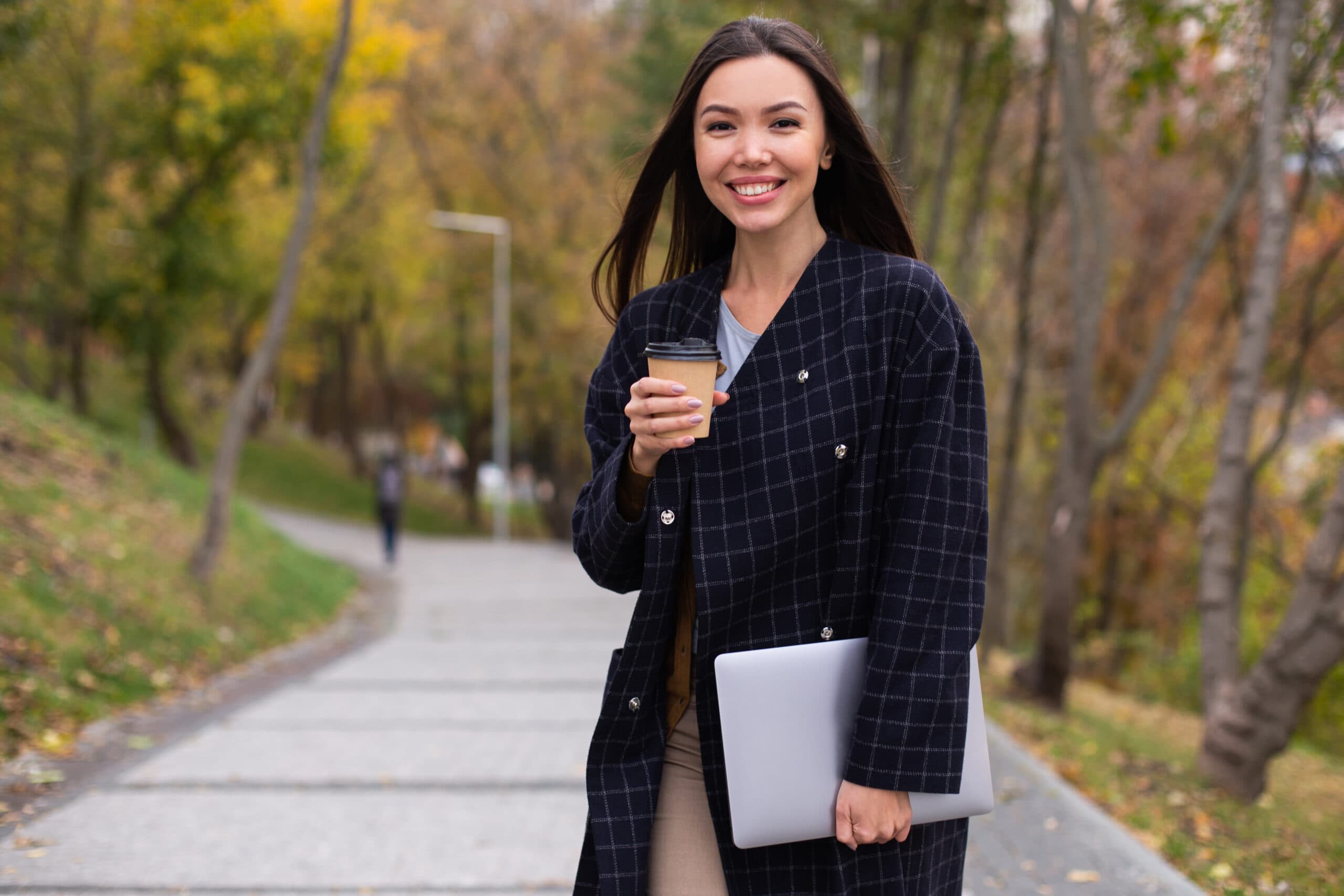 Young beautiful smiling woman in coat with coffee to go and lapt